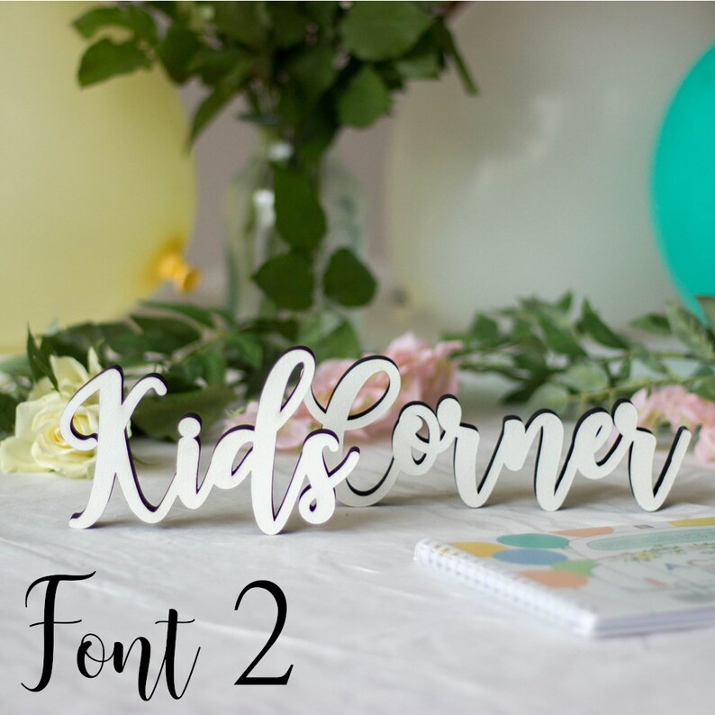 May include: White wooden sign with the text "Kids Corner" in a cursive font. The sign is on a white table with a notebook and flowers in the background. The text "Font 2" is written below the sign.
