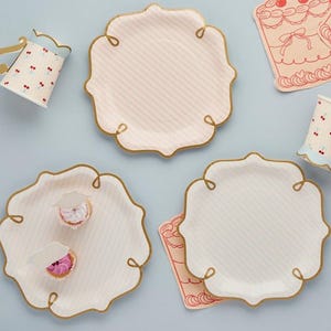 Puede incluir: Un conjunto de artículos para fiestas, que incluye platos con bordes festoneados, ribete dorado y un patrón de rayas. La imagen también muestra vasos a juego con diseños de cerezas y pequeños pastelitos en los platos. El tema general es una fiesta de té vintage.