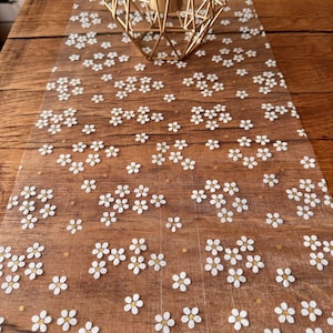 Op de afbeelding: Witte bloemen tafelrunner van een delicate, doorschijnende stof met een subtiele glans. De runner heeft een herhalend patroon van kleine witte bloemen op een transparante achtergrond.