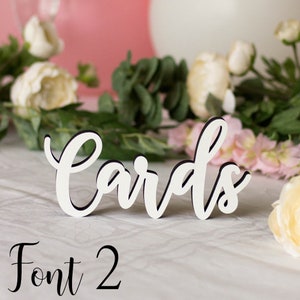 May include: White wooden sign with the word "Cards" written in a cursive font. The sign is sitting on a white table with flowers in the background. The text "Font 2" is written below the sign.