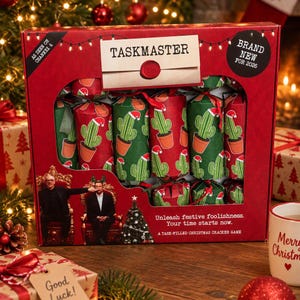 May include: A red rectangular box containing a Taskmaster Christmas cracker game. The packaging features a cactus pattern with Santa hats, the text "TASKMASTER," "AS SEEN ON CHANNEL 4," "BRAND NEW FOR 2026," and "Unleash festive foolishness. Your time starts now."