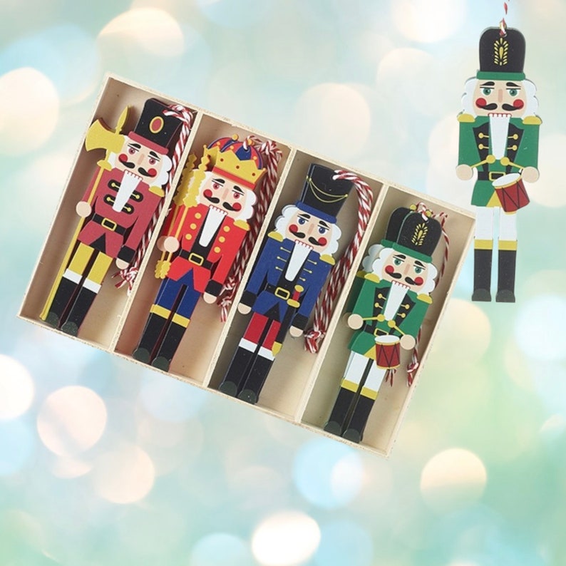 May include: A set of five flat wooden nutcracker ornaments in a partitioned box. The figures feature red, blue, and green uniforms with gold accents, black hats, and white beards. Each ornament includes a red and white twisted hanging string.