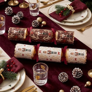 May include: Two Christmas crackers featuring nutcracker illustrations, one with a dark brown background and one with a white background, both tied with dark red ribbons. The crackers are arranged on a deep red velvet table runner with pinecones.