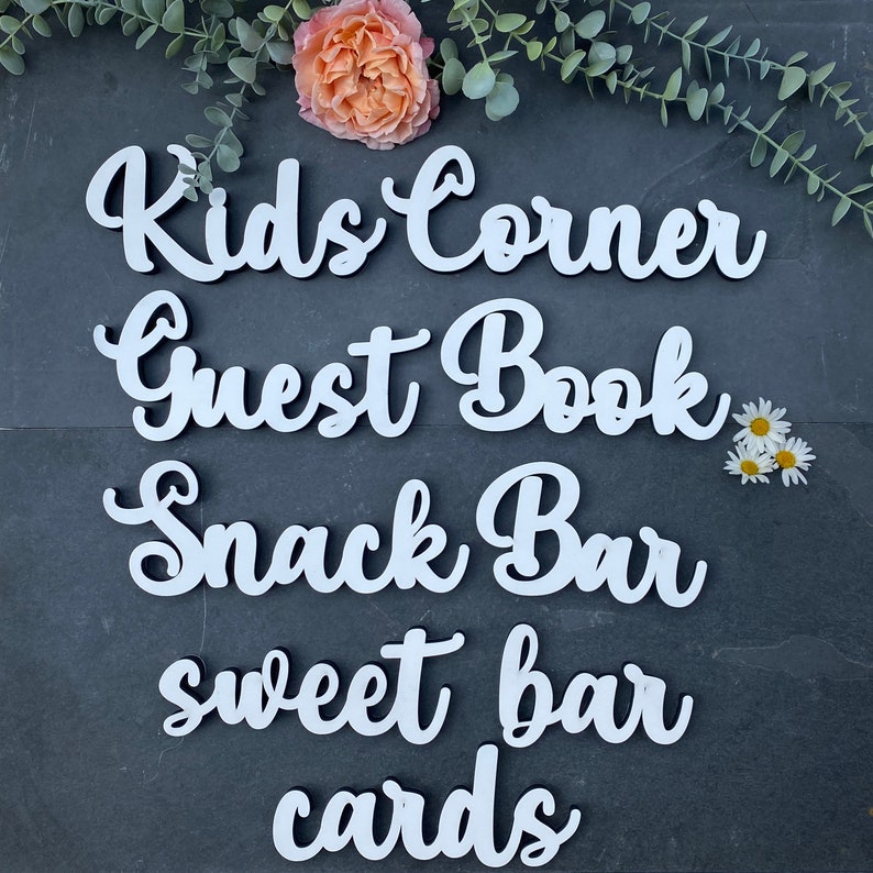 May include: White wooden signs with the words "Kids Corner", "Guest Book", "Snack Bar", "Sweet Bar", and "Cards" written in a cursive font. The signs are arranged in a vertical stack on a dark gray background.
