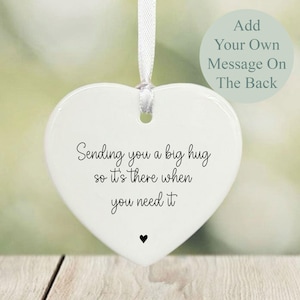 Pode incluir: Enfeite de cerâmica branco em forma de coração com uma fita branca. O enfeite tem o texto "Sending you a big hug so it's there when you need it" em escrita preta. Um pequeno coração preto na parte inferior.