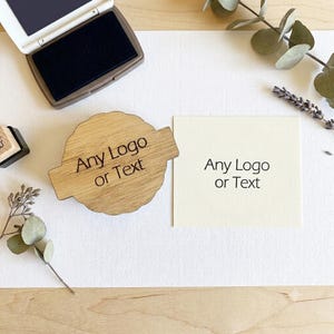 以下が含まれることがあります： Any Logo or Textという文字が刻印された持ち手付きの丸い木製スタンプと、黒いインクパッド、そして中央に同じくAny Logo or Textと黒いインクで印刷されたオフホワイトの正方形の紙が配置されています。