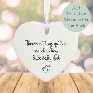 May include: White ceramic heart-shaped ornament with the text "There's nothing quite so sweet as tiny little baby feet" and a pair of footprints. The back of the ornament is blank and can be personalized with a message.