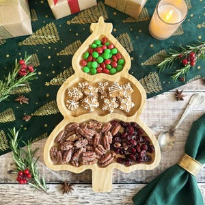 Peut inclure: Un plateau de service en bois clair en forme de sapin de Noël avec trois sections distinctes. La section supérieure contient des bonbons verts et rouges, la section centrale des biscuits en forme de flocon et la base des noix de pécan et des canneberges.