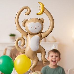 May include: A large, tan monkey-shaped balloon holding a yellow banana. The balloon has a white belly and face. Two other balloons, one green and one yellow, are at the bottom. A child in a green shirt and party hat smiles in the background.