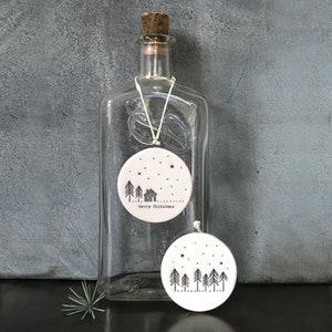 May include: Two white ceramic Christmas decorations with black and white illustrations. One decoration features a house and trees with the text "Merry Christmas". The other decoration features a row of trees.