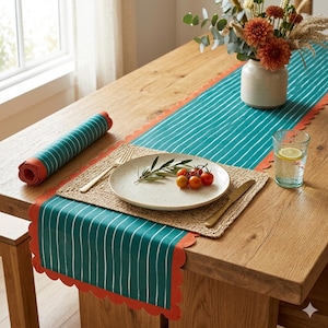 May include: A dining table set with a teal and white striped table runner and matching rolled placemat. The runner has a scalloped orange border. A woven placemat, plate, and gold cutlery are set for a meal. A glass of water and a vase of flowers complete the scene.