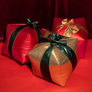Op de afbeelding: Drie cadeau dozen in rood en goud, met zwarte en gouden linten en strikken. De dozen zijn geplaatst op een rode ondergrond tegen een donkerrode achtergrond. De cadeaus hebben een glanzende, metallic uitstraling.