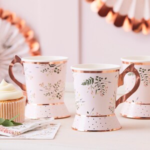 K&ouml;nnte beinhalten: Drei rosa Papierbecher mit Blumenmuster und goldenen Akzenten. Die Becher haben einen goldenen Henkel und einen goldenen Punktboden.