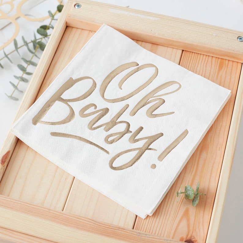 May include: White paper napkin with gold foil lettering that says "Oh Baby!" The napkin is sitting in a wooden tray.