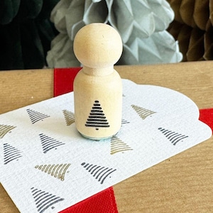 May include: A wooden stamp featuring a black and white geometric Christmas tree design. The stamp is mounted on a wooden handle and is used to create a repeating pattern of Christmas trees.