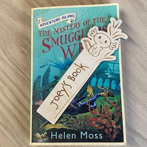 May include: A wooden bookmark featuring a hand-drawn illustration of an axolotl. The bookmark is personalised with the name "Joey's Book" and is resting on a book titled "The Mystery of the Smugglers' Wreck" by Helen Moss.