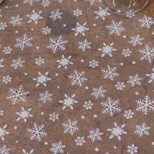 May include: A close-up of a brown fabric featuring a repeating pattern of white snowflakes and small white dots. The snowflakes are of varying sizes and designs, creating a winter holiday theme.