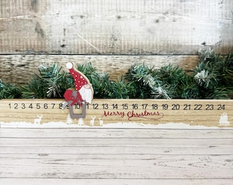 Może przedstawiać: Drewniany kalendarz adwentowy z numerami od 1 do 24. Zawiera figurkę gnoma i napis "Merry Christmas" w czerwonym kolorze. Zawiera małe wycięcia reniferów i drzew. Zieleń umieszczona za kalendarzem.