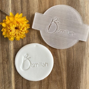 May include: A white plastic stamp with a handle and a round surface. The stamp has the word "Bismillah" written in a cursive font.