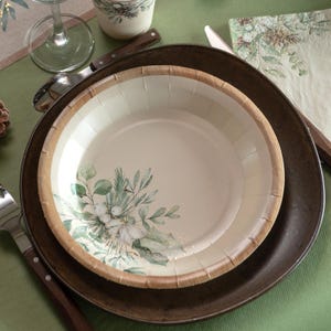 Puede incluir: Un plato de papel color crema con un diseño floral con hojas verdes y flores blancas, sobre un plato marrón oscuro. El plato tiene un borde de grano de madera natural. Se ven un tenedor y una cuchara.