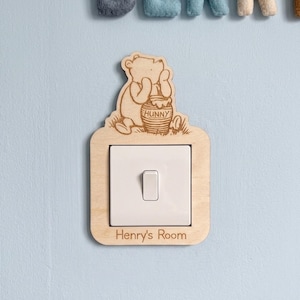 Könnte beinhalten: Holz-Lichtschalterabdeckung mit einer geschnitzten Abbildung von Winnie Puuh, der einen Honigtopf mit dem Wort "Hunny" hält. Die Abdeckung ist rechteckig mit dem Text "Henry's Room" am unteren Rand. Der Schalter ist weiß.