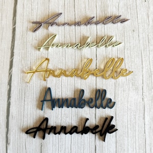 May include: Five cursive name cutouts spelling "Annabelle" in various colors: lavender, light green, gold, navy blue, and black. The names are displayed on a light wood-grain surface.