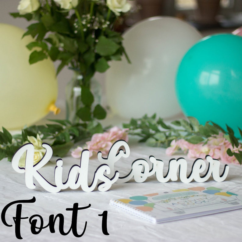 May include: White wooden sign with the words "Kids Corner" in a cursive font. The sign is on a white table with a notebook and flowers.