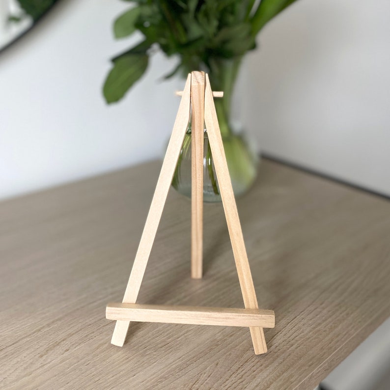 May include: A wooden easel with a clear glass vase on top. The vase is empty and the easel is made of natural wood.