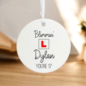 May include: A white, round ceramic ornament with the text "Blimmin' L Dylan YOU'RE 17" in black script. The letter "L" is inside a red square. A white ribbon is attached for hanging.