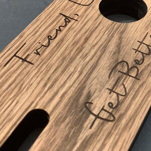 May include: Wooden drink holder with the words "Friend, I Get Better" etched into the surface. The holder has two circular cutouts and a long, rectangular slot. The wood grain is visible.