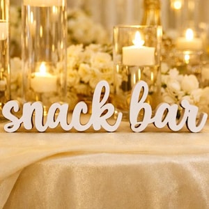 May include: A white wooden "snack bar" sign in a cursive font, set on a gold tablecloth. The background includes lit candles in glass holders and white flowers, creating a warm atmosphere for a party or event.