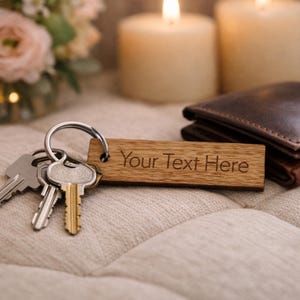 May include: A wooden rectangular keychain with the engraved text "Your Text Here" is attached to a silver keyring with several keys. A brown leather wallet is in the background, along with lit candles and flowers.