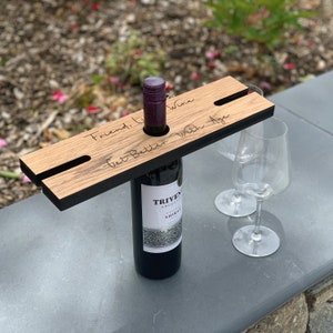 May include: A wooden wine bottle holder with a black border, holding a bottle of red wine. The holder has two slots for wine glasses and the engraved text reads "Friend, Like Wine, Get Better With Age."