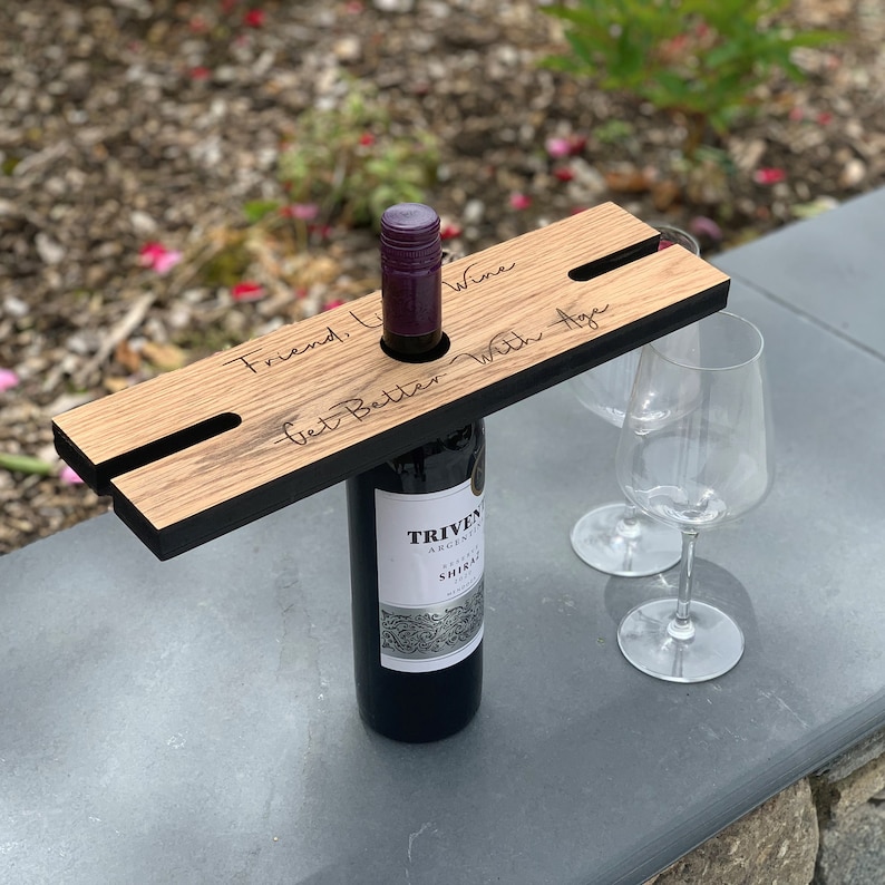 May include: A wooden wine bottle holder with a black border, holding a bottle of red wine. The holder has two slots for wine glasses and the engraved text reads "Friend, Like Wine, Get Better With Age."
