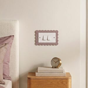 May include: A mauve-coloured light switch cover with a scalloped edge, featuring two white switches. A small gold-coloured analogue clock and two books are on a wooden bedside table next to a bed.