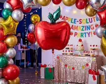 Puede incluir: Un gran globo de aluminio con forma de manzana roja y una hoja verde, suspendido frente a una decoración de fiesta con un cartel que dice APPLE-SOLUTELY AMAZING PARTY, un pastel de varios niveles y un arco de globos metálicos.