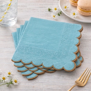 Op de afbeelding: Een stapel lichtblauwe papieren servetten met een decoratieve rand en een geschulpte rand. De servetten hebben een textuur en een goudkleurige rand. De servetten zijn gestapeld op een wit houten oppervlak.