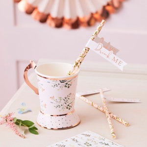 K&ouml;nnte beinhalten: Ein Set aus rosa und gr&uuml;nen floralen Papierstrohhalmen mit einer wei&szlig;en Flagge, auf der in goldenen Buchstaben "Drink Me" steht. Die Strohhalme befinden sich in einem rosa und gr&uuml;nen floralen Papierbecher mit goldfarbenem Rand und Henkel.