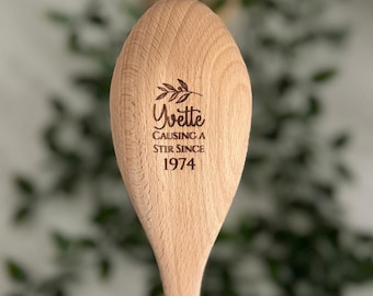 Puede incluir: Una cuchara de madera grabada con el texto "Yvette Causing a Stir Since 1974". La cuchara tiene un acabado de madera natural y un pequeño diseño de rama de olivo.