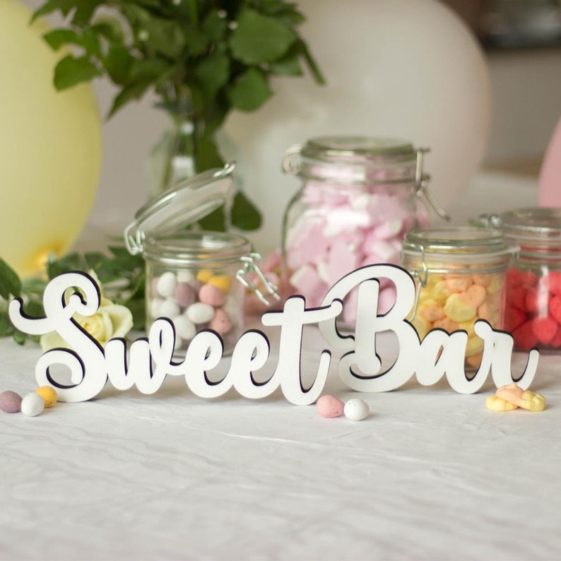 Puede incluir: Un letrero de madera blanco con las palabras "Sweet Bar" en una fuente cursiva. El letrero est&aacute; sobre un mantel blanco con tarros de dulces en el fondo.