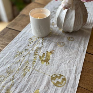 Op de afbeelding: Een witte linnen tafelrunner met een gouden folieontwerp van takken, konijnen en paaseieren. Een witte kaars en een witte keramische vaas met roze bloemen staan op de tafelrunner.