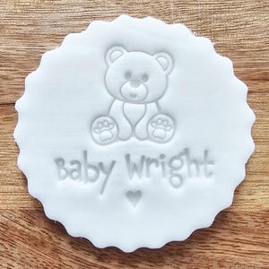 May include: A white, scalloped-edge cookie cutter with a teddy bear design and the words "Baby Wright" embossed on it. A small heart is below the text. The cutter is on a wooden surface.