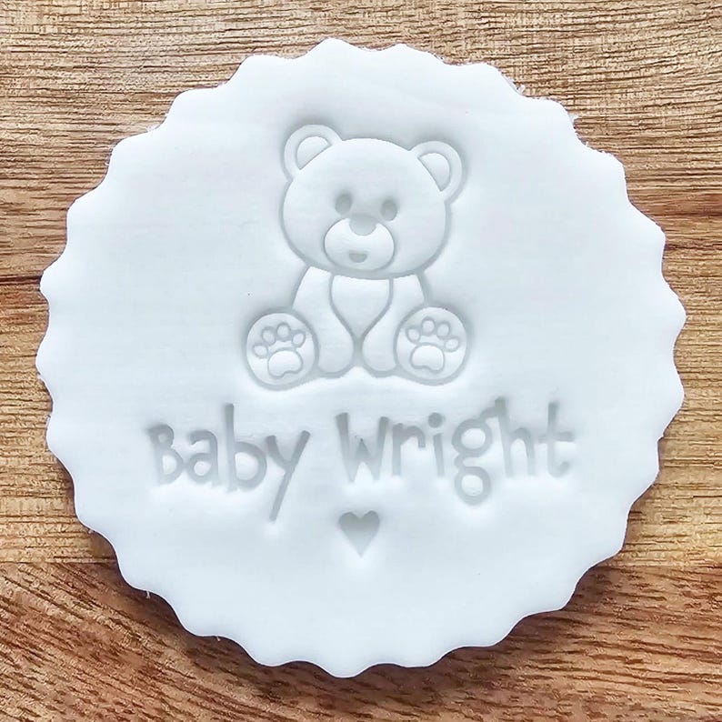 May include: A white, scalloped-edge cookie cutter with a teddy bear design and the words "Baby Wright" embossed on it. A small heart is below the text. The cutter is on a wooden surface.