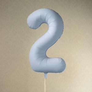 Könnte beinhalten: Hellblauer Folienballon in Form der Zahl 2. Der aufgeblasene Ballon hat eine matte Oberfläche. Er hängt an einem cremefarbenen Band vor einem strukturierten Hintergrund. Ideal für Geburtstage.