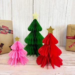 Könnte beinhalten: Drei Weihnachtsbäume aus Papier in Rosa, Grün und Rot, jeder mit einem goldenen Glitzerstern gekrönt. Die Bäume haben ein Wabenmuster und stehen vor einem hellen Holzhintergrund. Geschenkverpackte Geschenke sind sichtbar.