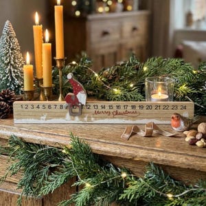 May include: A wooden Christmas advent calendar block featuring numbers 1 through 24, the text Merry Christmas, and a small movable gnome figure. The light wood piece displays white silhouettes of deer and trees along the bottom edge.