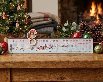 Könnte beinhalten: Ein rechteckiger hölzerner Weihnachts-Countdown-Kalender mit einer Winterdorf-Illustration, den Zahlen 1 bis 25 und einem beweglichen Wichtel-Marker. Der Text lautet Ellie's Christmas Countdown. Das Design zeigt verschneite Landschaften, Häuser und Bäume.