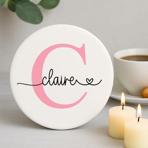 Może przedstawiać: Okrągła biała podstawka ceramiczna z różową literą "C" i imieniem "claire" w kursywie. Małe serce następuje po imieniu. Dwie zapalone świece i filiżanka kawy w tle.