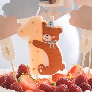 May include: A number one candle with a brown bear design, wearing a blue party hat, and holding the number. The candle is decorated with small gold stars. The candle is on a cake with strawberries and raspberries.