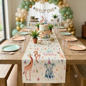 May include: A table runner featuring illustrations of a giraffe, monkey, and elephant wearing party hats, with the text "HAPPY BIRTHDAY". The table is set with light green plates, clear glasses, and a two-tier cake topped with animal figurines.